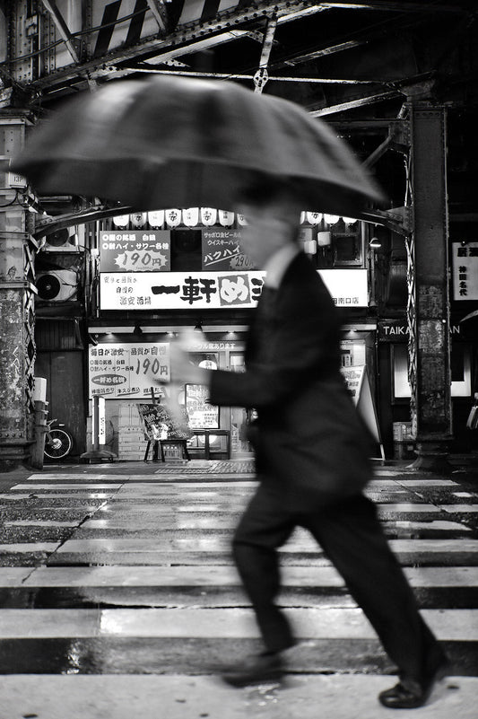 A black and white image of a man walking in the rain with an umbrella near Kanda station
