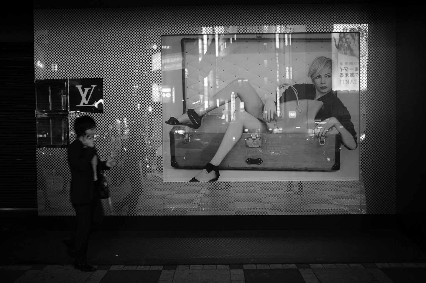A man walking on his phone walks past an Ikebukuro department store window display at night.