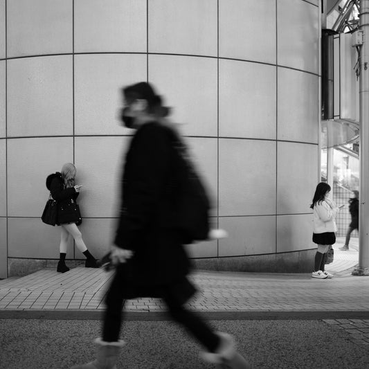 Two girls look at their phones while a woman rush past in a blur, Shibuya 109.