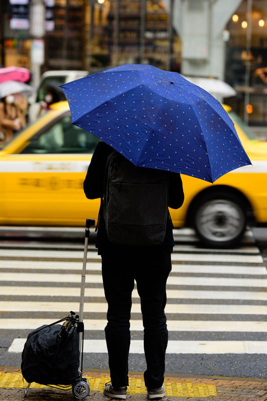 A man holds a blue umbrella as a yellow taxi passes by.