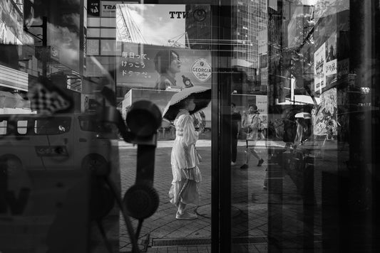 A woman with a sun umbrella walks through past a phone booth in Shibuya Crossing.