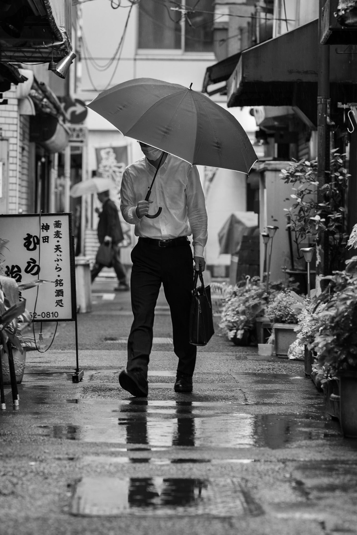 Two men walk in opposite directions in a rainy Kanda alley.