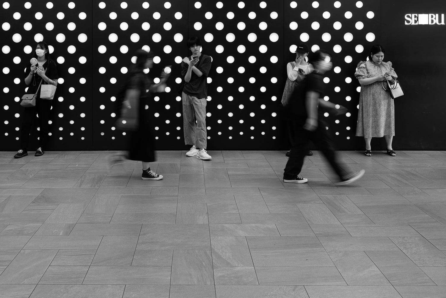 Four people wait outside Ikebukuro Seibu department store looking at their smartphones.