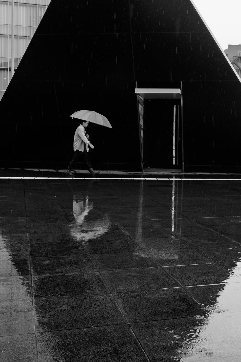 A black and white image of a man walking in the rain with an umbrella past a traingle-shaped entrance.  