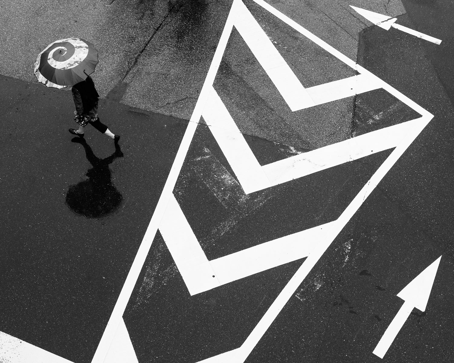 A black and white image of a woman walking through an intersection in the rain with a spiral umbrella.  Her reflection is seen on the road.