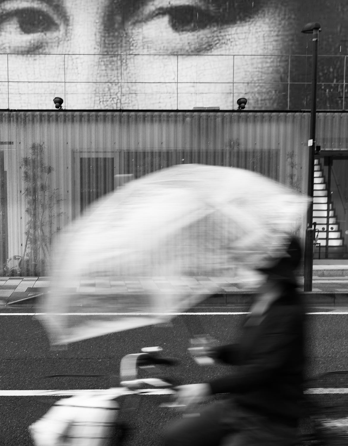 A woman ride a bicycle in the rain past a giant mona Lisa billboard.