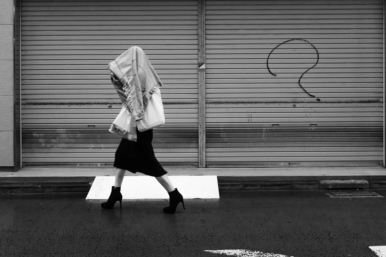 A black and white images of a woman walking in the rain with a jean jacket for an umbrella and high heel boots, past a spry painted question mark.