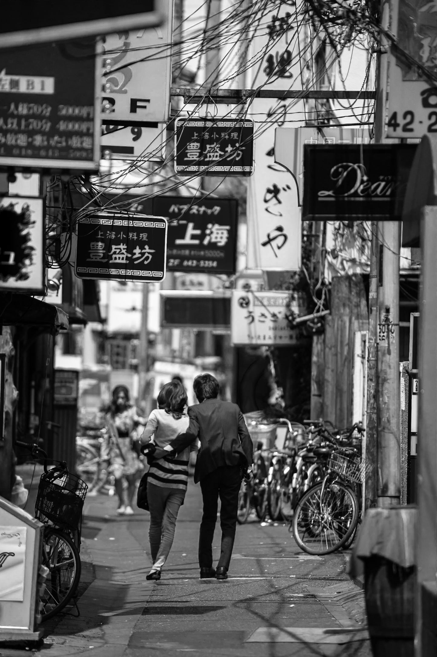 A black and white image of a couple walking at midday under the nightclub signage of Omiya.